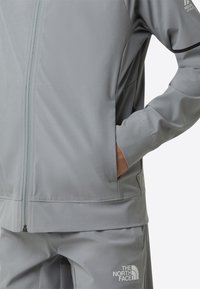 Lysegrå jakke med glidelåslukking, sidelommer og ribbestrikkede mansjetter. Har en diskret logo på brystet og matchende bukser nedenfor.