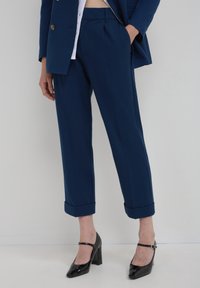 Pantaloni su misura blu navy con orlo risvoltato, dotati di tasche laterali e vita piatta, abbinati a scarpe col tacco nere lucide.
