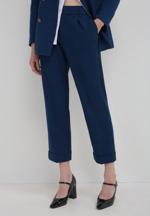 Marineblaue maßgeschneiderte Hose mit umgeschlagenem Saum, seitlichen Taschen und flacher Vorderseite, kombiniert mit glänzenden schwarzen High Heels.