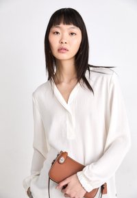 Blouse blanche à manches longues avec un décolleté en V et des poignets froncés ; tenant un petit sac en cuir marron avec des finitions en argent et une couture contrastante.