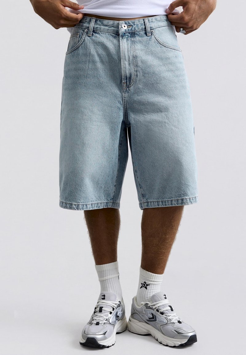 Homme portant un short en jean bleu clair, des chaussettes de sport blanches et des chaussures de course gris argenté, ajustant un t-shirt blanc.