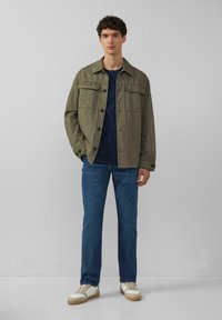 Olijfgroene jas met twee voorzakken, blauwe henley shirt eronder, blauwe jeans en witte sneakers met tan zolen.