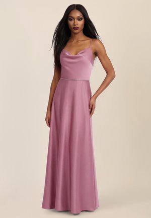 Kobieta o długich, prostych włosach ubrana w bezrękawnikową, liliową satynową suknię z cienkimi ramiączkami i ozdobnym pasem w talii, stojąca na beżowym tle.