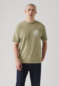 Olive green cotton t-shirt with a crew neck, featuring a white printed Levi's logo on the upper left chest area. Short sleeves and relaxed fit.
