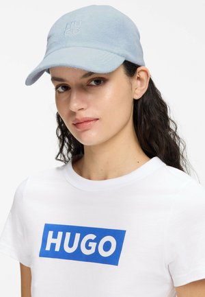Kvinde med langt mørkt hår iført en lyseblå kasket og hvid T-shirt med blåt HUGO-logo på hvid baggrund.