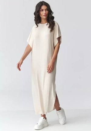 Kobieta o ciemnych, kręconych włosach, ubrana w luźną, jasnobeżową sukienkę maxi i białe sneakersy na platformie, stojąca na jednolitym tle.