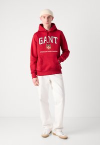 Sudadera roja con el logotipo "GANT" en blanco, con bolsillo tipo canguro, combinada con pantalones blancos y zapatillas crema sobre un fondo neutro.