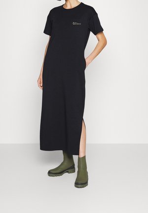 Kobieta w czarnej, krótkiej sukience midi z rozcięciem po bokach i kieszeniami, w połączeniu z oliwkowozielonymi botkami za kostkę.