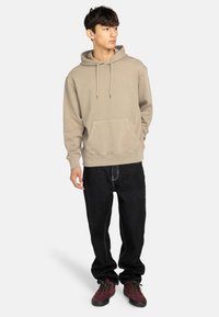 Beige huvtröja med ett framfick, kombinerad med svarta jeans och röd-burgundy skor. Har dragsnören och en avslappnad passform.