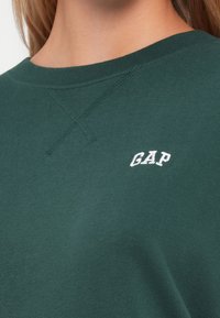 Felpa verde scuro con scollo a girocollo, polsini a costine e logo "GAP" ricamato sul petto. Tessuto di cotone liscio.
