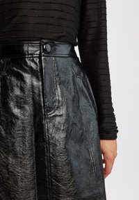 Fustă din piele neagră cu o textură lucioasă, buzunare laterale și închidere cu nasture. Asortată cu un top transparent negru, ajustat, cu mânecă lungă și dungi.