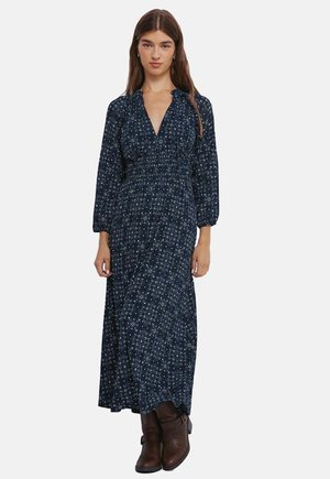 Navy blauwe maxi jurk met lange mouwen, v-hals, bloemenpatroon, gathered taille en vloeiende rok; gecombineerd met bruine enkellaarsjes.