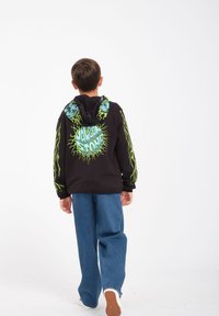 Czarna bluza z kapturem z zielonymi akcentami graficznymi i tekstem z tyłu. Niebieskie dżinsowe spodnie o luźnym kroju. Teksturowany materiał i swobodny design.