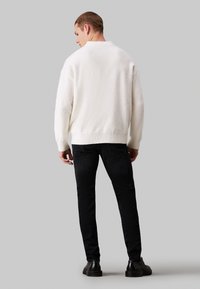 Suéter blanco texturizado con cuello redondo, dobladillo acanalado y ajuste holgado, combinado con jeans oscuros y calzado negro.
