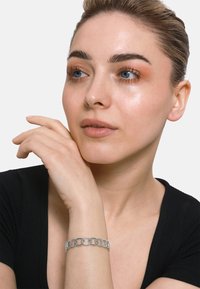 Silberne Gliederarmband mit kreisförmigen und strukturierten Elementen, getragen an einem hellhäutigen Arm. Der Hintergrund ist schlicht weiß, und das Modell hat dezentes Makeup.