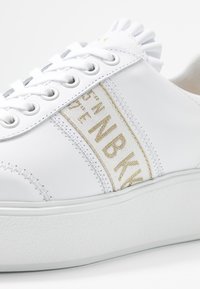 Bílé kožené tenisky s bílými tkaničkami, silnou podešví, zlatým prošíváním a béžovým textilním panýlkem s textovým detailem po straně.