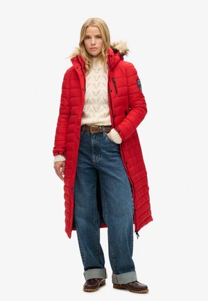 Red padded long coat with a fur-lined hood, zippered pockets, and a quilted texture. Worn over a cream sweater and blue wide-leg jeans.