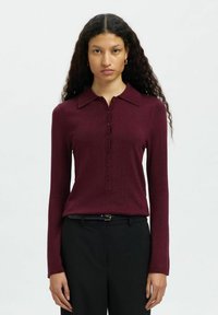 Pikkade lokkis juustega naine kannab tumepunast nuppudega pikkade varrukatega pluusi ja musti pükse musta vööga, seistes ühetoalise heletausta ees.