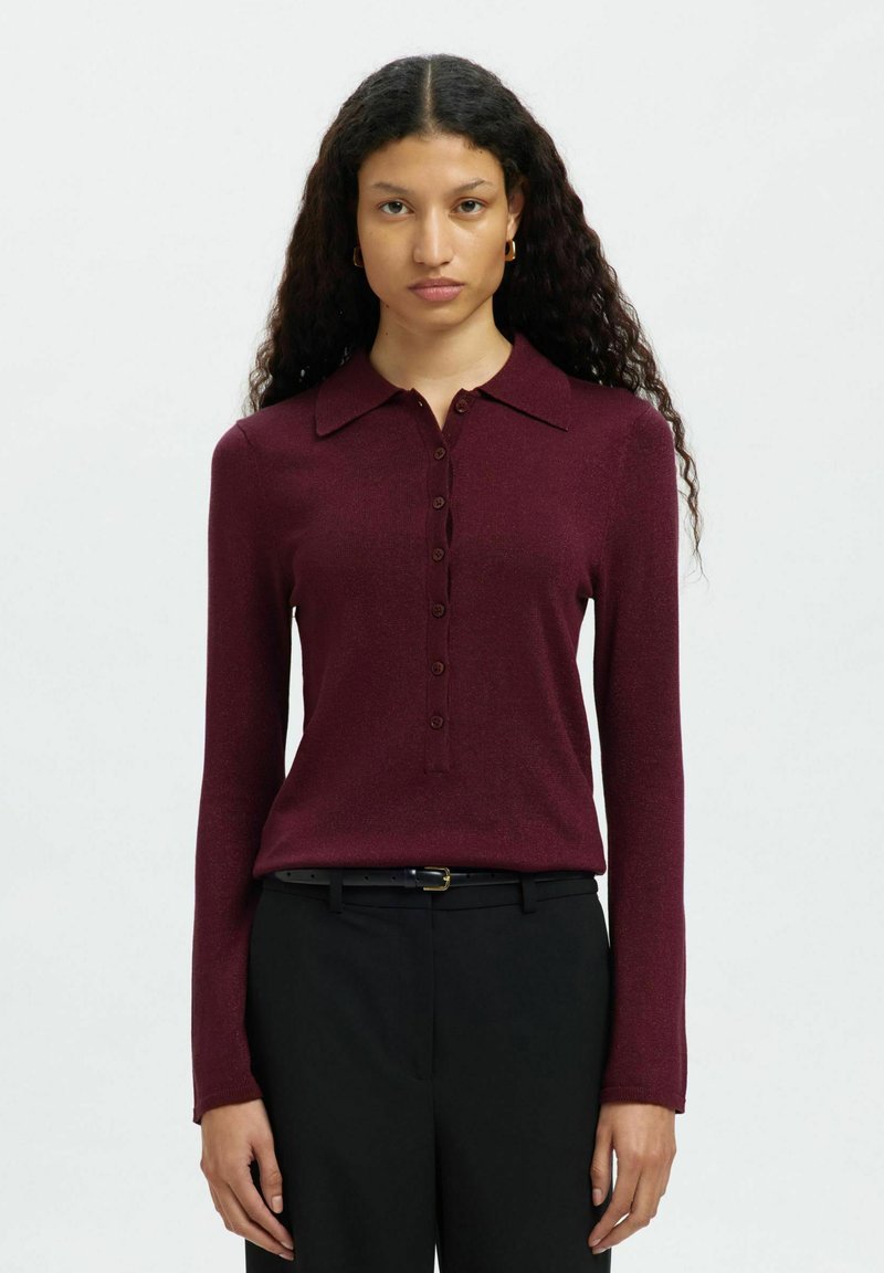 Pikkade lokkis juustega naine kannab tumepunast nuppudega pikkade varrukatega pluusi ja musti pükse musta vööga, seistes ühetoalise heletausta ees.