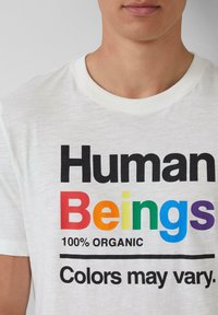 Nahaufnahme eines weißen T-Shirts mit dem Text "Menschen" in Regenbogenfarben, "100% BIO" und "Farben können variieren" unter dem Text.