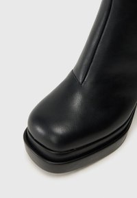 Pogržden posnetek črnih usnjenih čevljev s kvadratnim prstom in debelim podplatom na enostavni svetli podlagi.