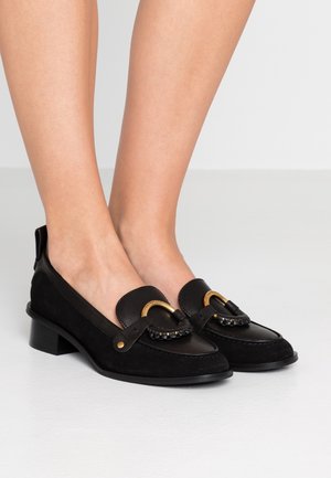 Mocassins noirs à talon bas avec boucles décoratives dorées portés pieds nus sur un fond clair uni.