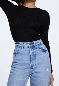 Mujer vistiendo una camiseta de manga larga negra con canalé metida dentro de unos vaqueros ajustados de tiro alto, azules claros y desgastados, posando con una mano detrás de la cabeza.