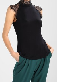 Top negro sin mangas con un alto cuello de encaje y acentos de encaje en los hombros, hecho de un material suave; combinado con pantalones verdes holgados.