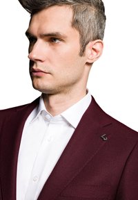 Burgundowa marynarka o teksturowym wzorze z wyrazistym kołnierzem, połączona z elegancką białą koszulą, ozdobiona czarną przypinką na klapie.