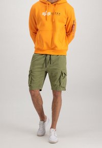 Homme portant un sweat à capuche orange Alpha Industries, un short cargo vert olive et des baskets blanches, debout devant un fond clair uni.