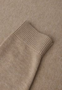 Le tissu tricoté beige présente un bord de manche en côtes. La texture est lisse avec une légère variation de couleur. Dans l'ensemble, il a une apparence douce.