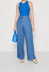 Blå ärmlös topp med logotyp, vida denimjeans och en liten blå handväska. Klumpiga vita sandaler med detaljer. Släta texturer och avslappnad design.