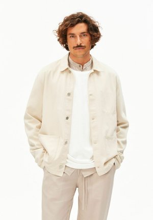 Homme aux cheveux bouclés et moustache portant une veste beige, une chemise blanche et un pantalon beige, debout les mains dans les poches sur fond blanc.
