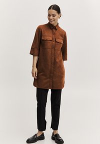 Braun, samtiges Hemdkleid mit durchgehendem Knopfverschluss, zwei Brusttaschen und Dreiviertelärmeln, kombiniert mit schwarzen maßgeschneiderten Hosen und Mokassins.
