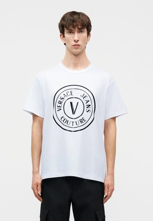 Νεαρός άνδρας με κοντά καστανά μαλλιά, φορώντας λευκό μπλουζάκι Versace Jeans Couture και μαύρο παντελόνι cargo, με απλό ανοιχτό φόντο.