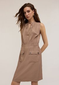 Rochie fără mâneci, de culoare beje, cu un gât strâns, talie elastică și două buzunare frontale cu detalii cu nasturi. Țesătură moale și texturată.
