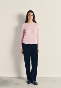 Moteris stovi priešais paprastą sieną, dėvinti šviesiai rožinį ilgomis rankovėmis megztinį, tamsiai mėlynas korduro kelnės ir tamsiai mėlynas austi batus.