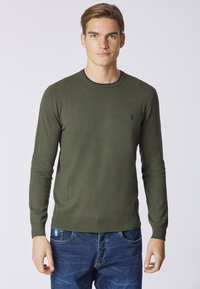 Maglione verde lavorato a maglia con scollo rotondo, maniche lunghe e logo ricamato sul petto. Abbinato a jeans in denim blu.