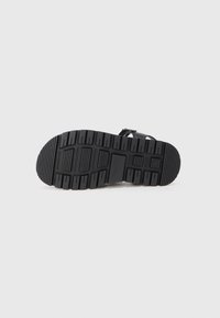 Crna sandala leži na boku, prikazujući debeli, žlijebasti potplat s grubim uzorkom gaznoga sloja na jednostavnoj svijetloj pozadini.