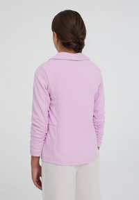 Top lungo a maniche con colletto in rosa chiaro, con maniche arricciate. Presenta una texture liscia e una vestibilità morbida, indossato su pantaloni beige.