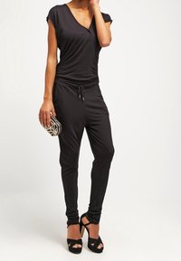 Svart jumpsuit med korta ärmar, v-ringning och dragsko i midjan. Matchas med svarta klackskor med remmar och en svart mönstrad clutch.