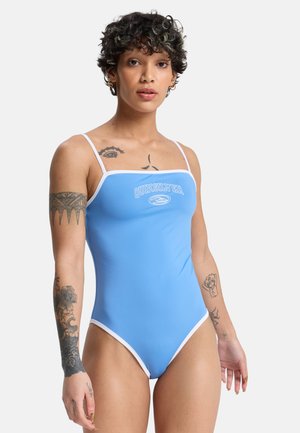 Junge Person mit kurzem lockigem Haar, die einen blauen Badeanzug mit weißem Saum und Quiksilver-Logo trägt und mehrere Arm- und Brusttattoos zeigt.