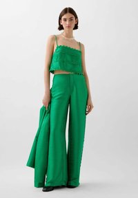Grøn cropped top med bølgede kanter, parret med højtaljede viddebukser i et struktureret stof. Modellen holder en matchende jakke.