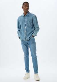 Camicia in denim azzurro chiaro con taschino, chiusura a bottoni e vestibilità rilassata, abbinata a jeans azzurro chiaro e sneakers bianco sporco.