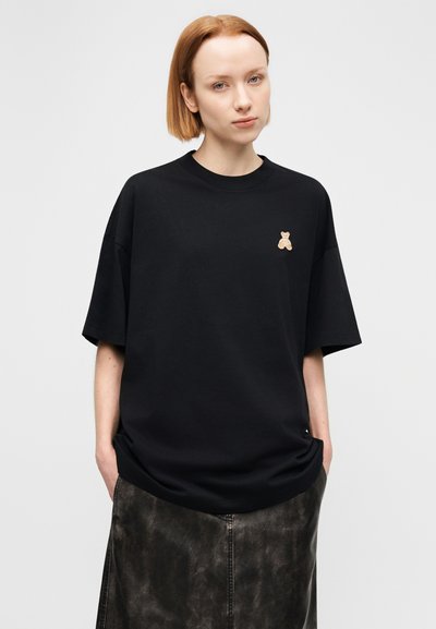 Μαύρο φαρδύ t-shirt από απαλό ύφασμα με κοντά μανίκια, διακοσμημένο με μικρή κεντημένη αρκούδα σε ταμπά χρώμα στο αριστερό στήθος. Η υφή της φούστας είναι από τεχνητό δέρμα.