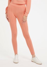 Leggings de couleur pêche avec une taille haute, présentant des coutures visibles et une texture lisse, associés à des baskets blanches.