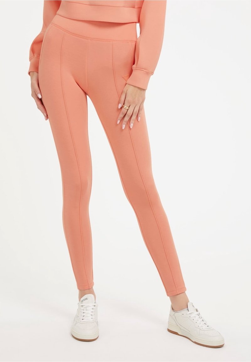 Leggings de couleur pêche avec une taille haute, présentant des coutures visibles et une texture lisse, associés à des baskets blanches.