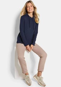 Felpa con cappuccio blu navy con lacci, abbinata a pantaloni beige chiaro e sneaker di colore chiaro, seduta su un piedistallo bianco.
