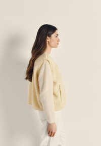 Šviesiai geltonas flisinis liemenėlis su minkštu, tekstūruotu paviršiumi ir viena šonine kišene, derinamas su baltomis kelnėmis. Švarios linijos ir laisvas siluetas.