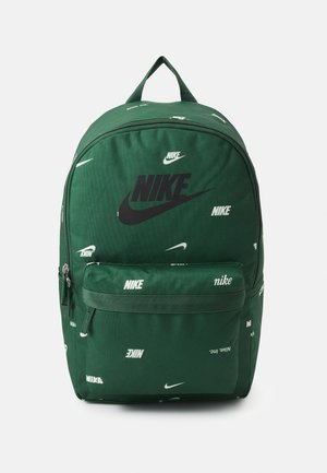 Mochila de tela verde con un destacado logo negro de Nike. Presenta varias marcas de Nike en blanco y un bolsillo frontal con cremallera. Asa acolchada en la parte superior.
